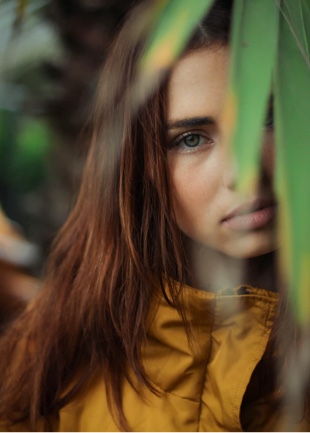 Girl in yellow jacket, with dangling leaves covering parts of her face.