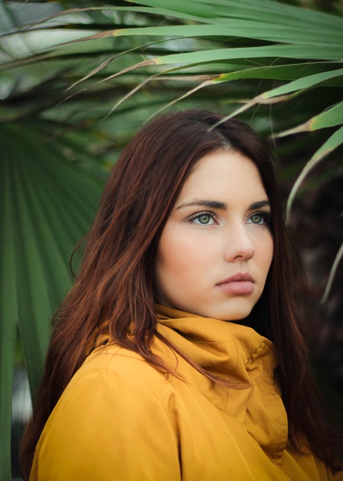 Girl in a yellow jacket, looking away from the viewer, surrounded by long tropical plant leaves.