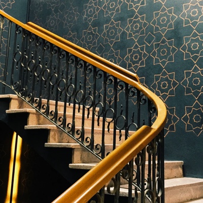 Curving wooden staircase with ornate, black metal balusters. Dark green wallpaper with gold accents in the background.